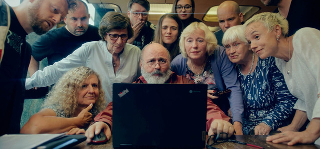 A group of older adults and a few younger people gathered around a laptop, looking intently at the screen.