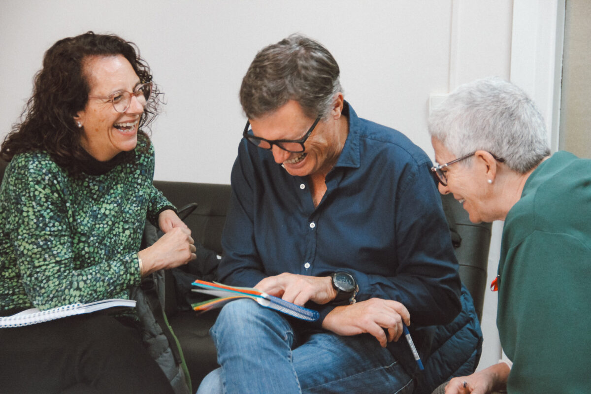 Three older adults, two women and one man, laughing together while looking at a notebook.