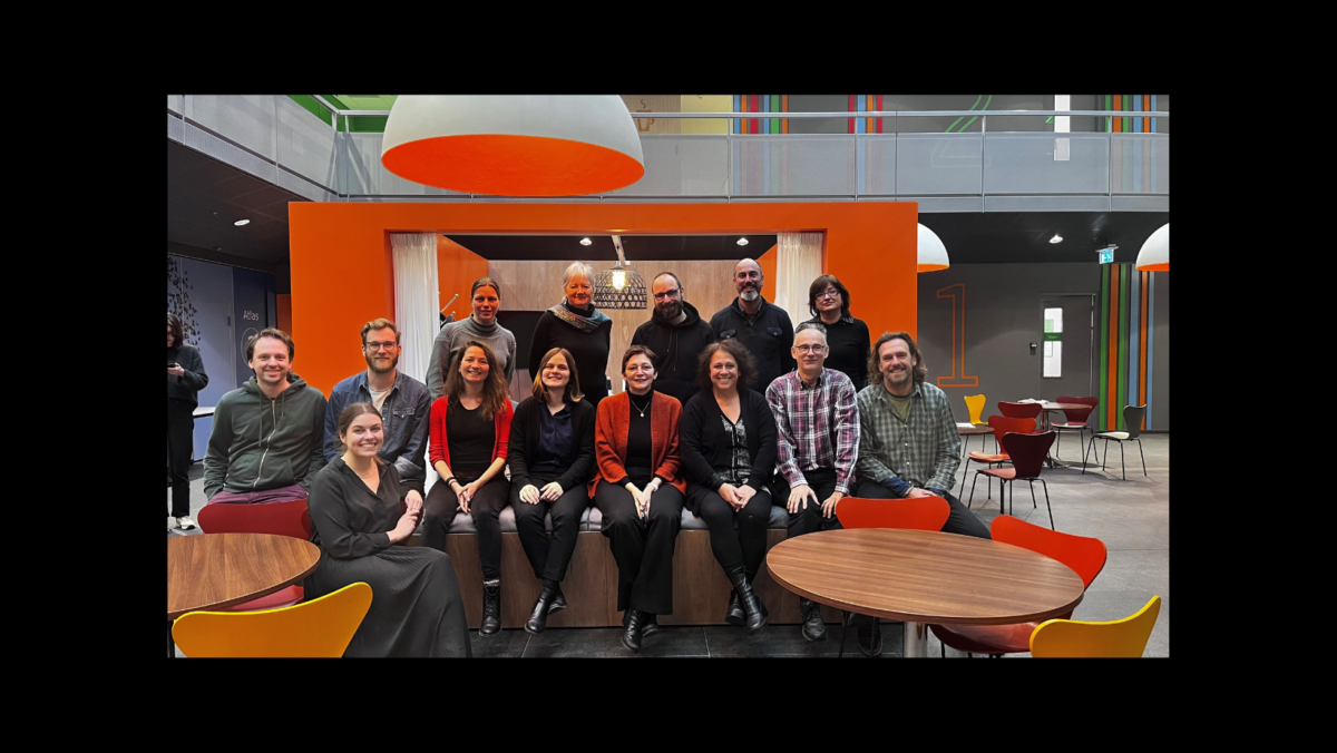 A group of 15 people, 10 women and 5 men, smiling and sitting together on a bench in an office.