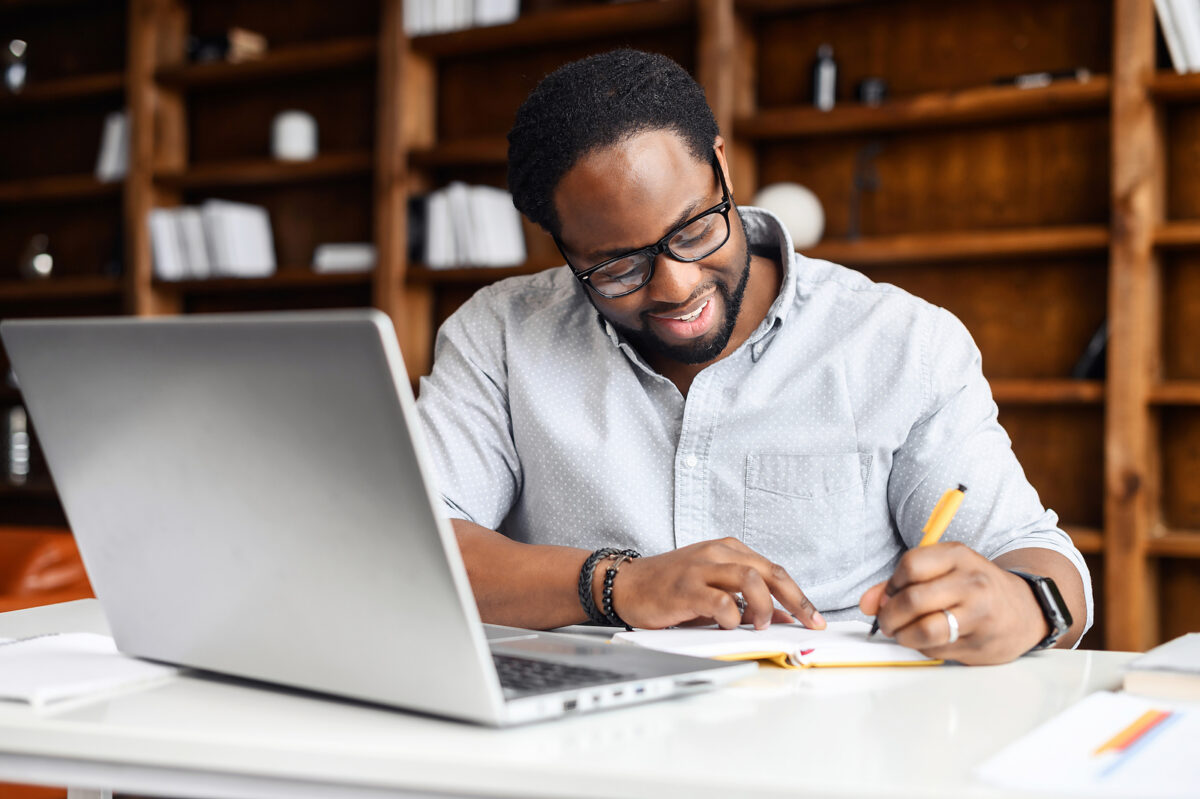 A smiling man in glasses and a light blue shirt writing in a notebook, with a laptop nearby.