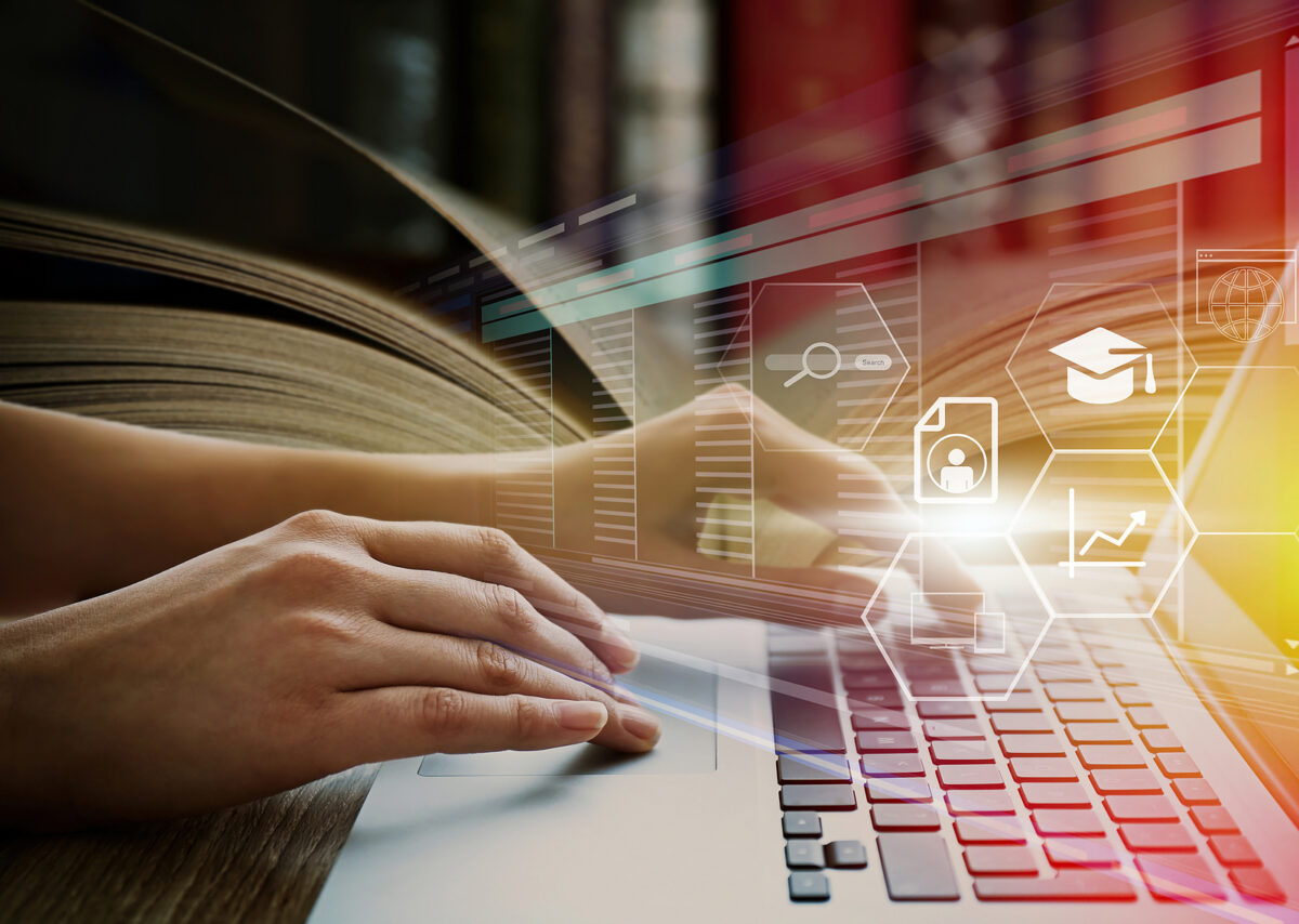 A person's hands typing on a laptop keyboard with a blurred open book in the background and digital icons overlaid.