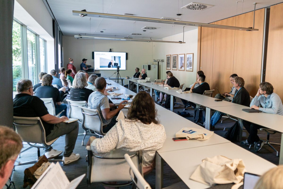 Attendees at a workshop, seated at tables, watching a presentation on a large screen.
