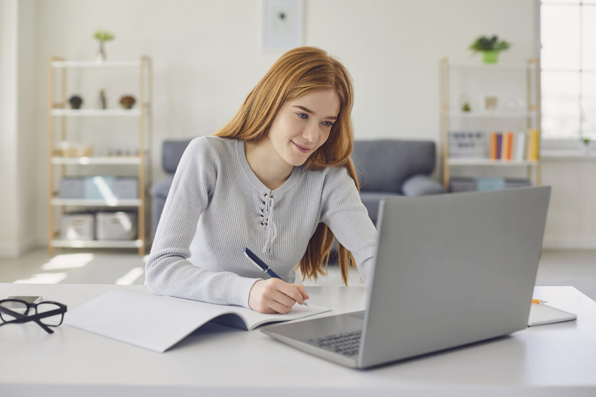 Smiling young woman with long red hair studying at a desk with a laptop and notebook
