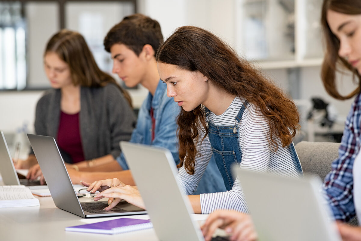 Students in a classroom working on laptops, focusing on a girl in a striped top and denim overalls.