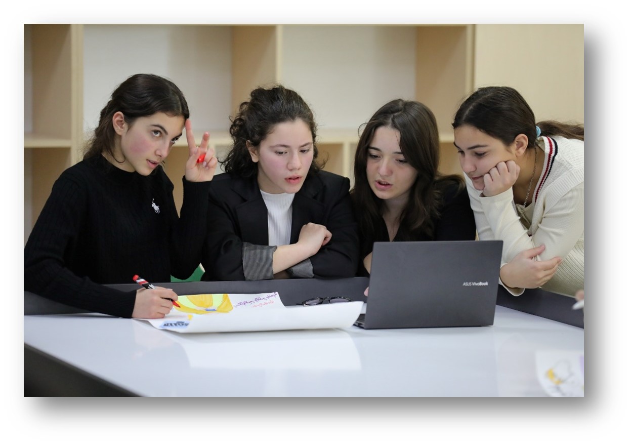 Four female students collaborating around a laptop and a drawing on a table, one making a peace sign.