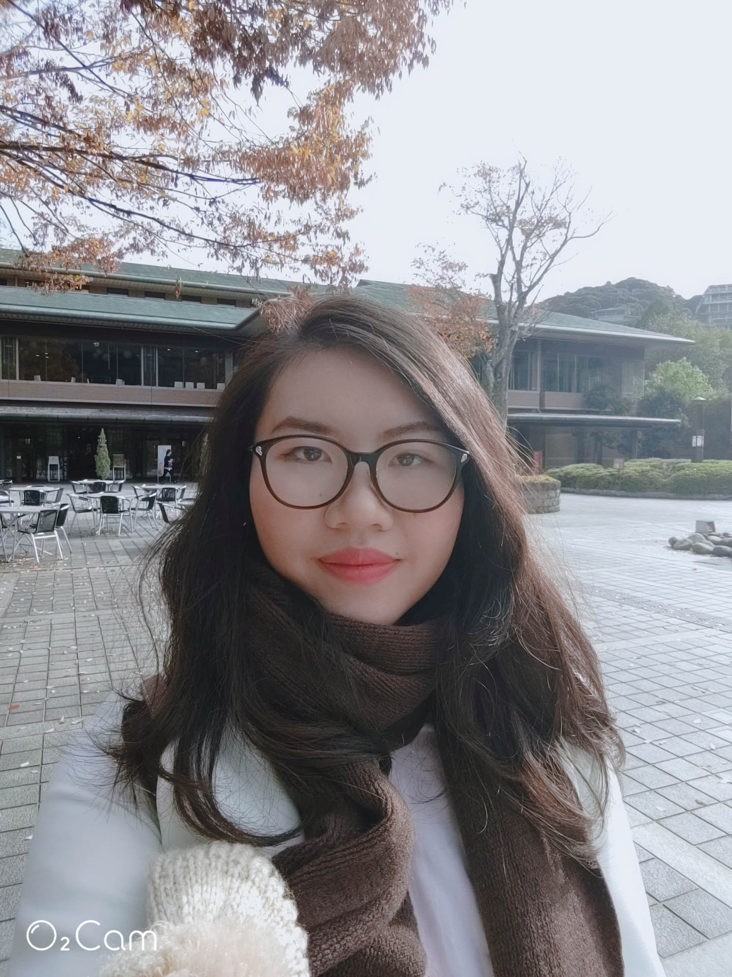 Zoe, a young woman with long dark hair and glasses, wearing a white top and brown scarf, smiling at the camera.