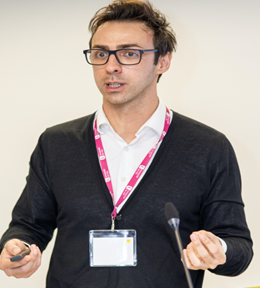 A speaker with dark hair and glasses presenting at a conference, holding a remote in one hand.