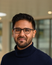 A man with dark hair and a beard, wearing glasses and a dark blue roll-neck jumper, smiles at the camera.