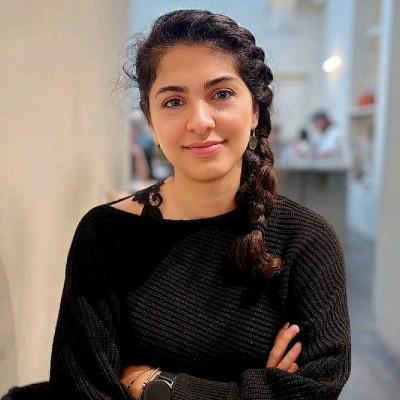 A young woman with dark braided hair and a black sweater smiles at the camera, arms crossed.