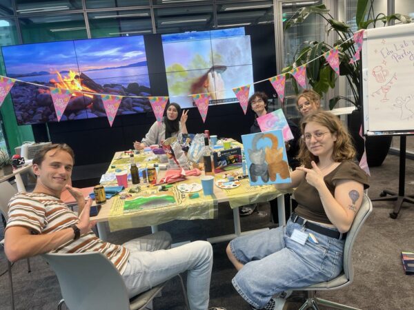 Five students painting at a table with party decorations, a whiteboard, and large screens in the background.